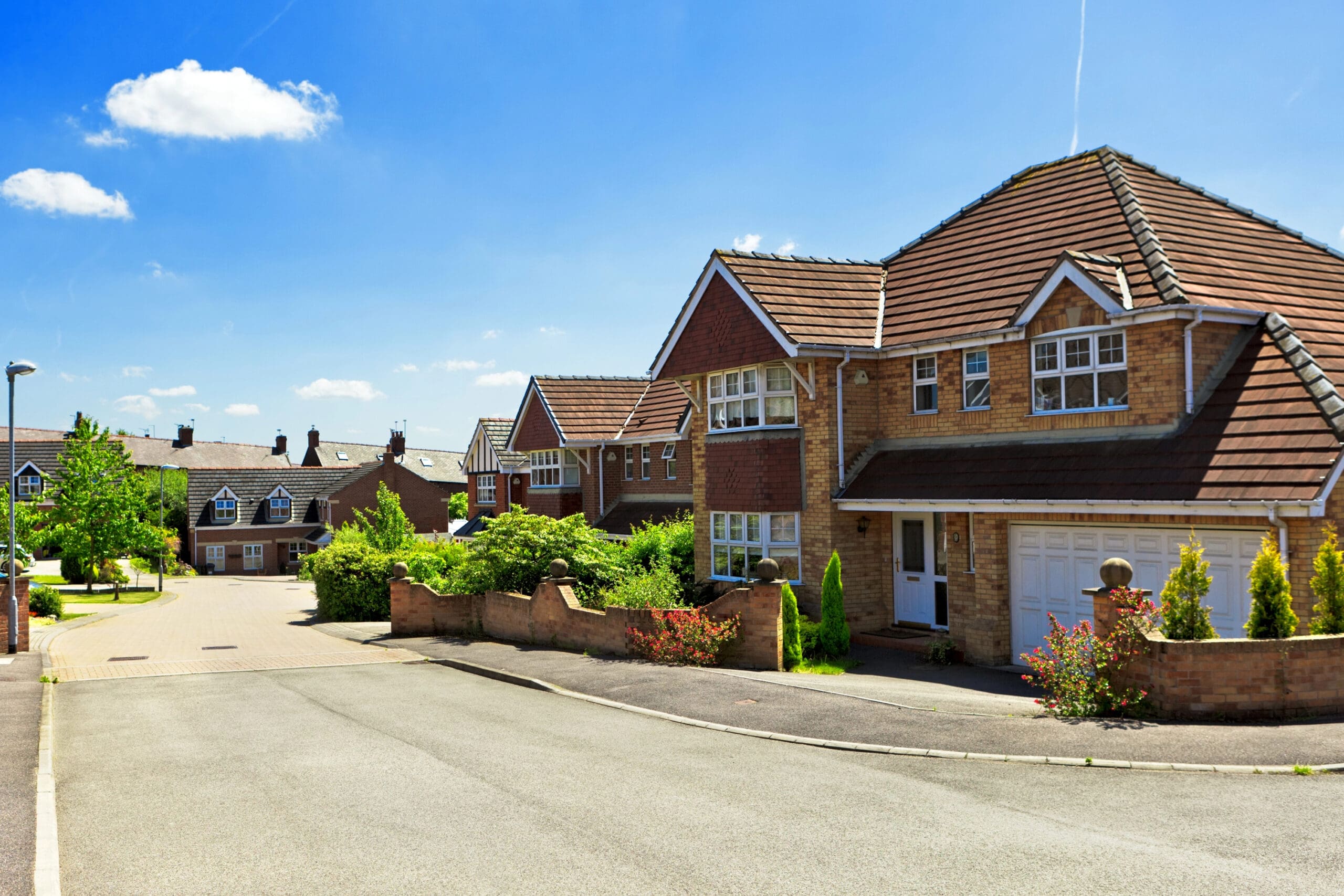 A residential street featuring modern, two-story brick houses with tiled roofs and well-maintained front gardens. The sky is clear and blue, indicating a sunny day. The street is quiet with no visible traffic or pedestrians.