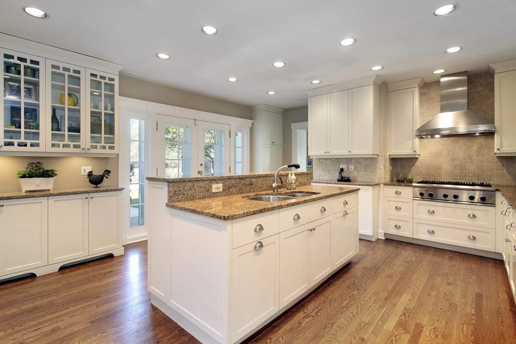 Spacious modern kitchen with white cabinetry, granite countertops, and stainless steel appliances. Central island with sink, wooden flooring, and glass-front cabinets. Recessed lighting illuminates the room. Double French doors open to the outside.