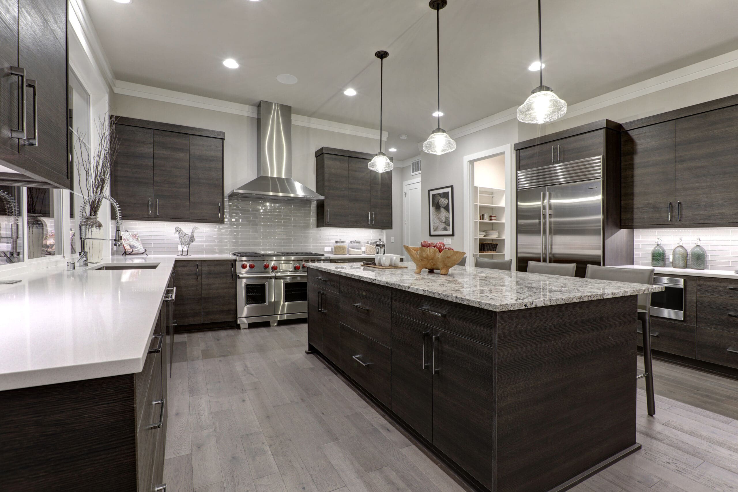 Modern kitchen with dark wooden cabinets, stainless steel appliances, and a large marble island with three hanging pendant lights. The room features a built-in refrigerator, a stove with a large hood, and a pantry in the background. The floor is light wood.