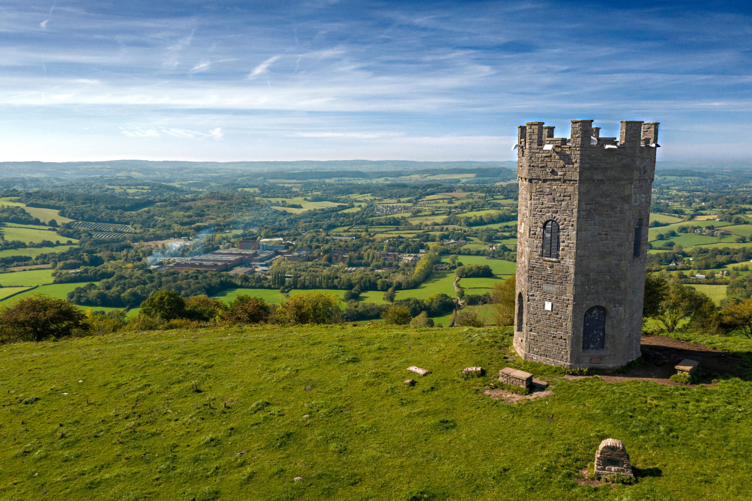 A stone tower stands on a grassy hill overlooking a vast, picturesque landscape. Rolling green fields, patches of woodland, and scattered buildings stretch into the distance under a clear blue sky with wispy clouds.