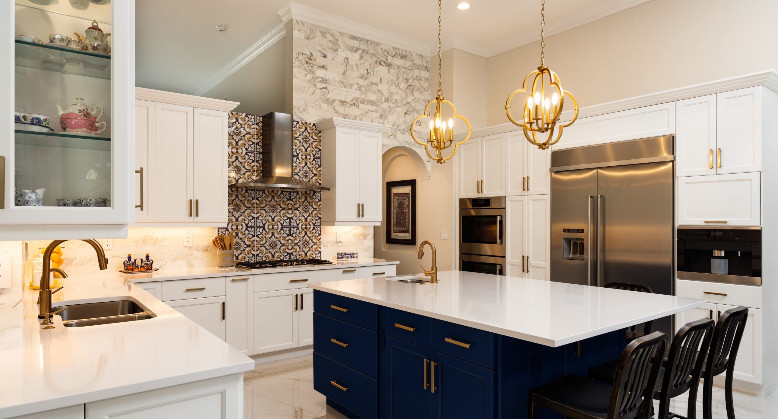 A modern kitchen featuring white cabinetry, a blue island with a white countertop, and stainless steel appliances. Two gold pendant lights hang above the island. The backsplash behind the stove showcases a decorative blue and white tile pattern.