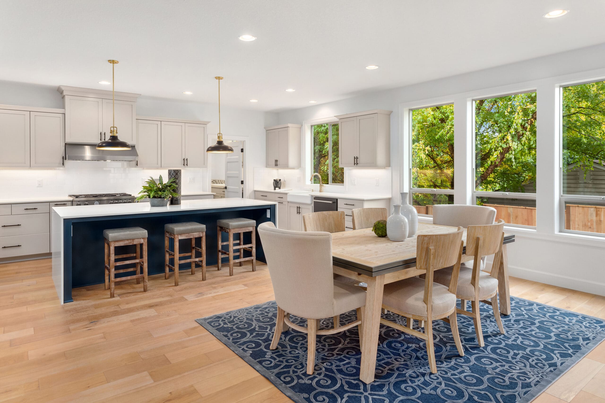 A modern kitchen and dining area featuring light-colored cabinetry, stainless steel appliances, and a large island with stools. A wooden dining table with mixed seating is on a blue rug. Large windows offer a view of green trees outside, and the space is well-lit.