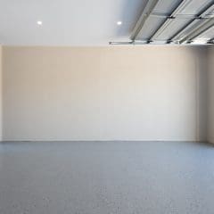 A spacious, empty garage with a clean, gray, speckled floor. The garage door is partially open, revealing a glimpse of the outdoors. A tall sliding glass door is situated on the left side, and a small plant decorates the right corner outside the garage.