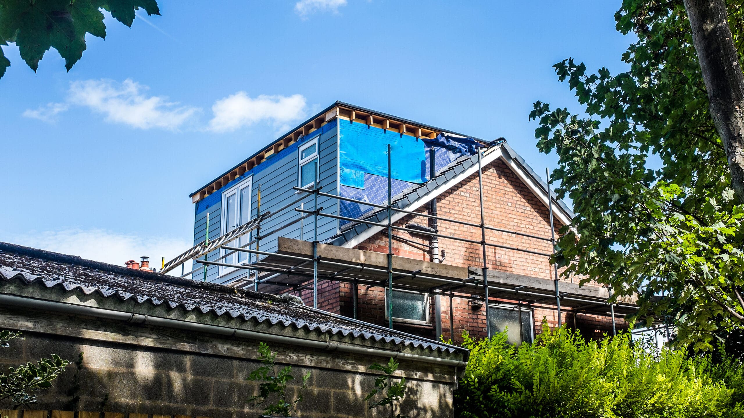 A house under construction with scaffolding on one side. The upper part of the house is partially covered with blue tarps. Surrounding the house are green trees and shrubs under a clear blue sky.