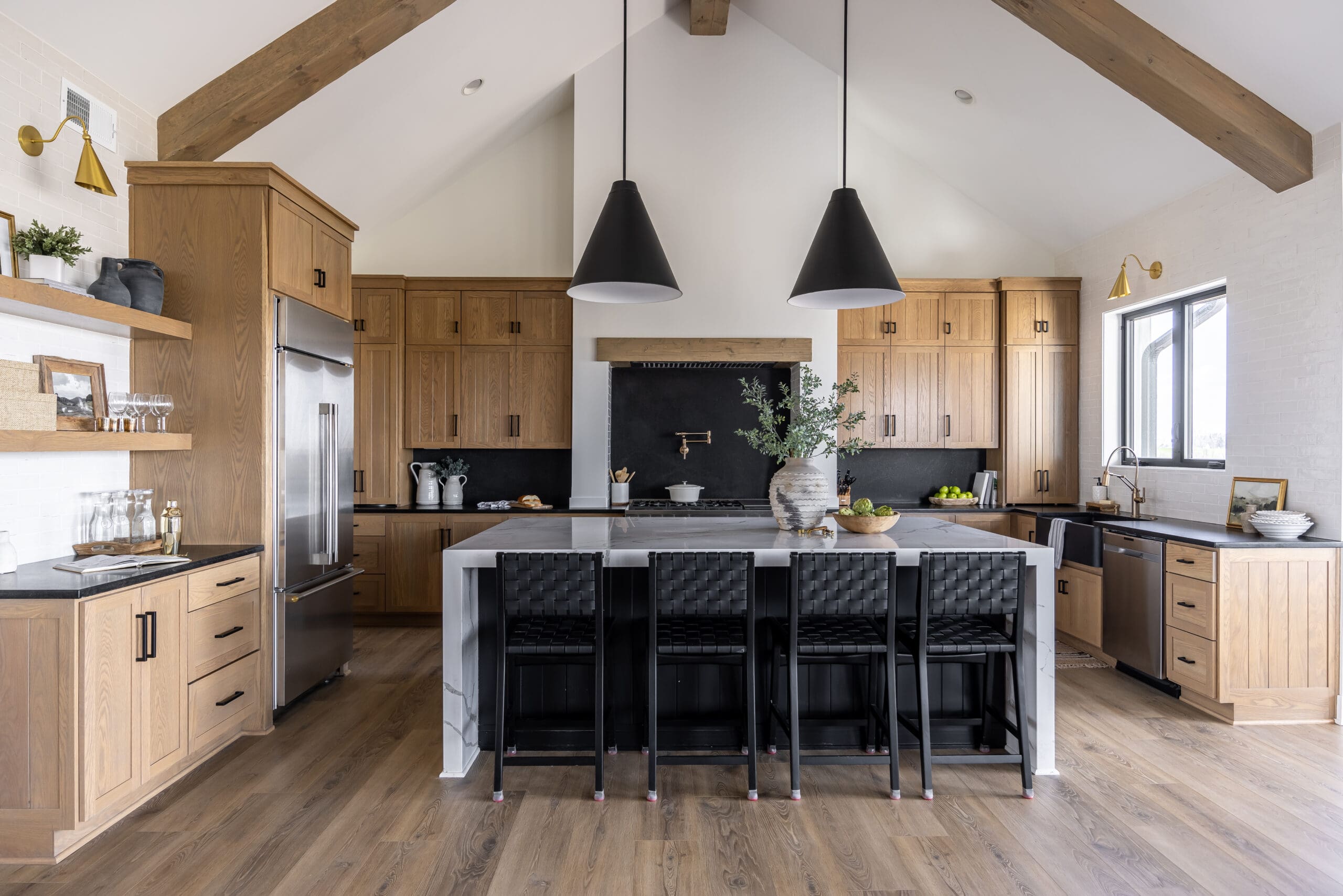 A modern kitchen with wooden cabinets, a large island with a marble countertop, and four black woven chairs. The kitchen features black pendant lights, stainless steel appliances, and exposed wooden beam ceilings. Light streams in through a window above the sink.