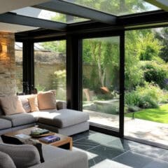 A cozy living room with a large gray sectional sofa adorned with soft cushions on a tiled floor. Floor-to-ceiling glass doors open to a lush garden with a manicured lawn, trees, and plants, allowing natural light to fill the space. A wooden coffee table holds decor items.