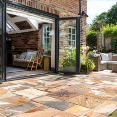 A sunlit patio area with stone tiles features a wicker-style seating arrangement with cushions. Tall glass doors open to reveal an indoor space with a rocking chair and various furnishings. The patio is surrounded by greenery and flowers in vibrant bloom.