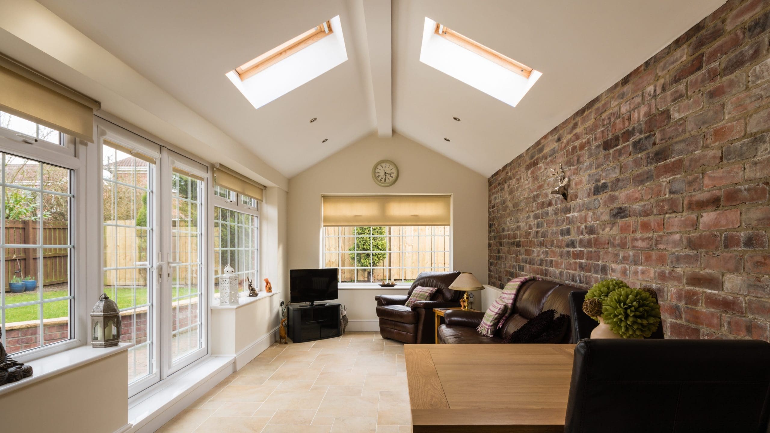 A bright sunroom with skylights and large windows. The room has a brick wall on the right, two leather armchairs with a small table between them, a TV on a stand, and a wooden dining table with chairs. The room overlooks a garden with a wooden fence.