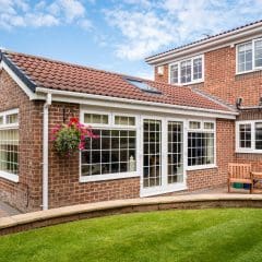 A modern brick house with large windows and a red-tiled roof. Attached is a sunroom with glass doors. The backyard features a manicured lawn, a patio with hanging flower baskets, and wooden benches near the entrance. Blue sky with some clouds above.