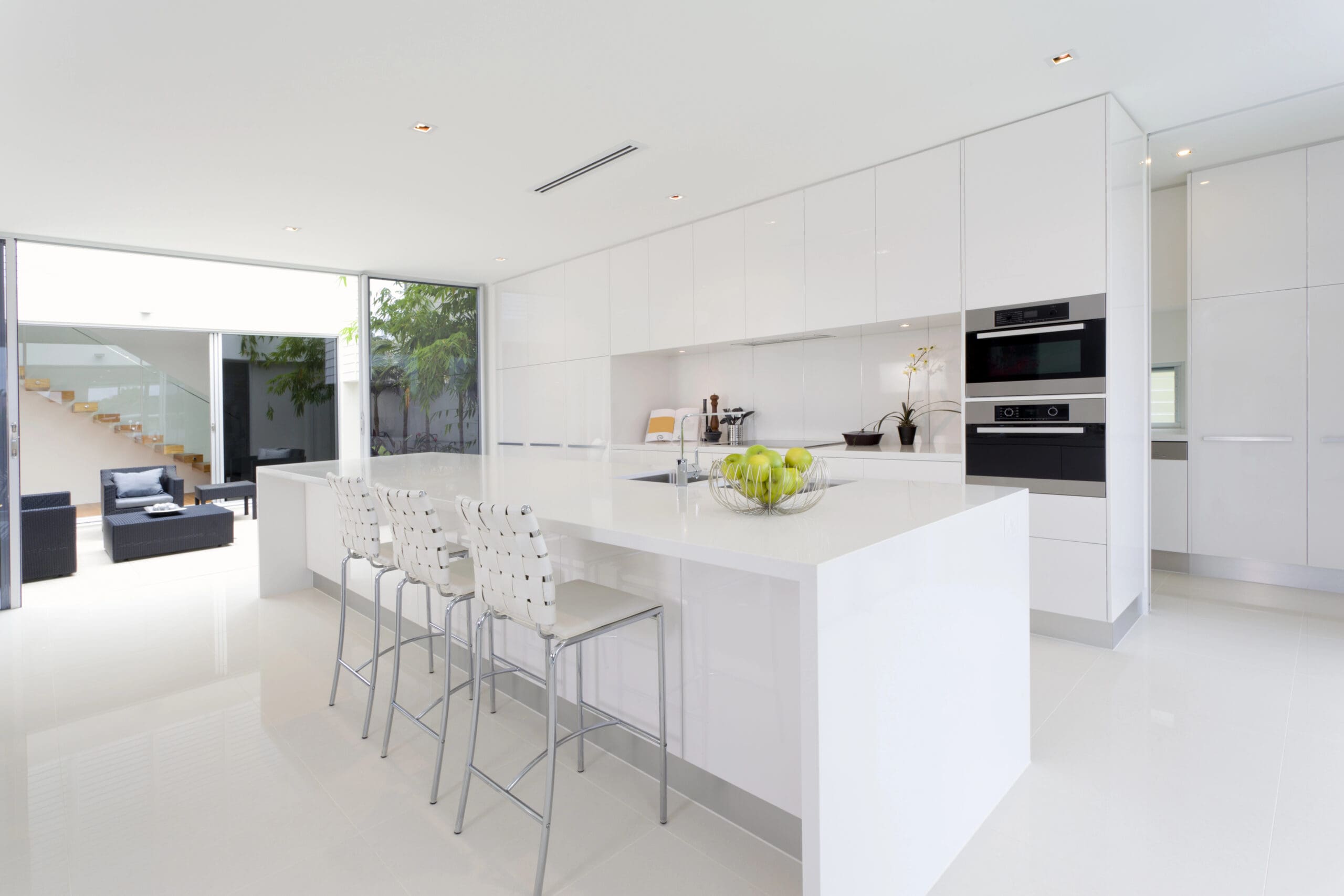 A modern kitchen with white cabinets and countertops. There is a large island with four high stools, a bowl of green apples on the island, and built-in appliances. The kitchen opens to a living area in the background, which features a staircase and sofas.