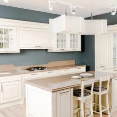 A modern, spacious kitchen with white cabinetry and beige countertops. It features a large island with two chairs and pendant lighting, as well as a built-in stove, oven, and glass-fronted cabinets. The walls are painted a soft blue-gray.