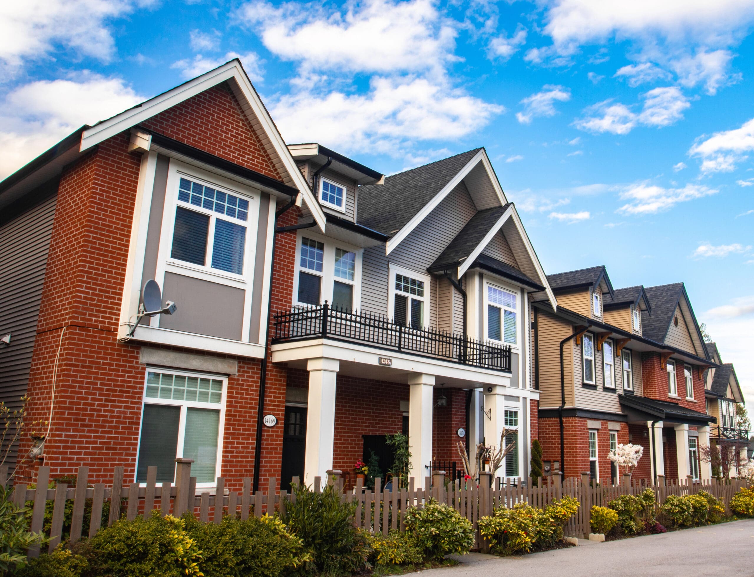 Row of modern, two-story houses with varied exteriors of brick and siding. Each home features large windows, gabled roofs, and small front yards with shrubs. A wooden fence runs along the property line. The sky is partly cloudy.