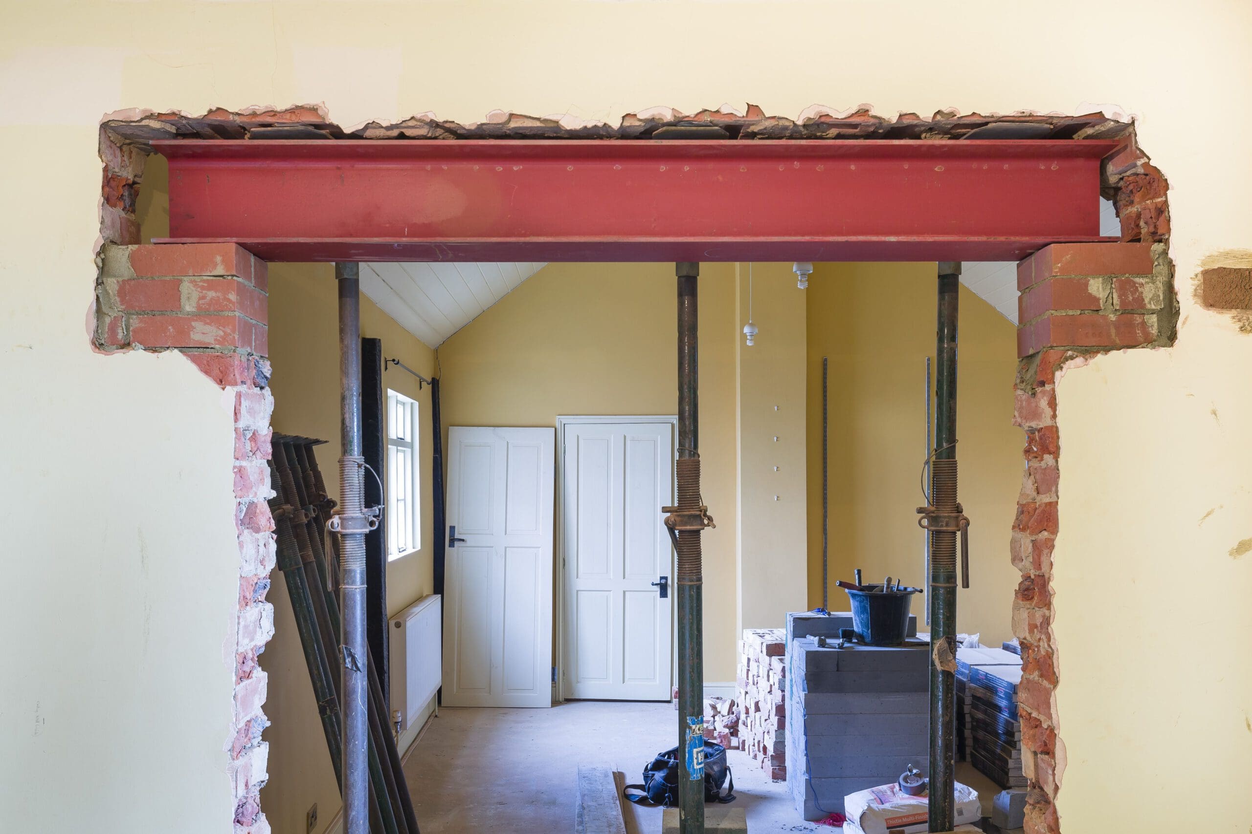 An unfinished room under construction with a steel beam installed across a partially demolished wall. The floor has various construction tools and materials, including support beams and a stack of bricks. The room has a window and double doors on the opposite wall.