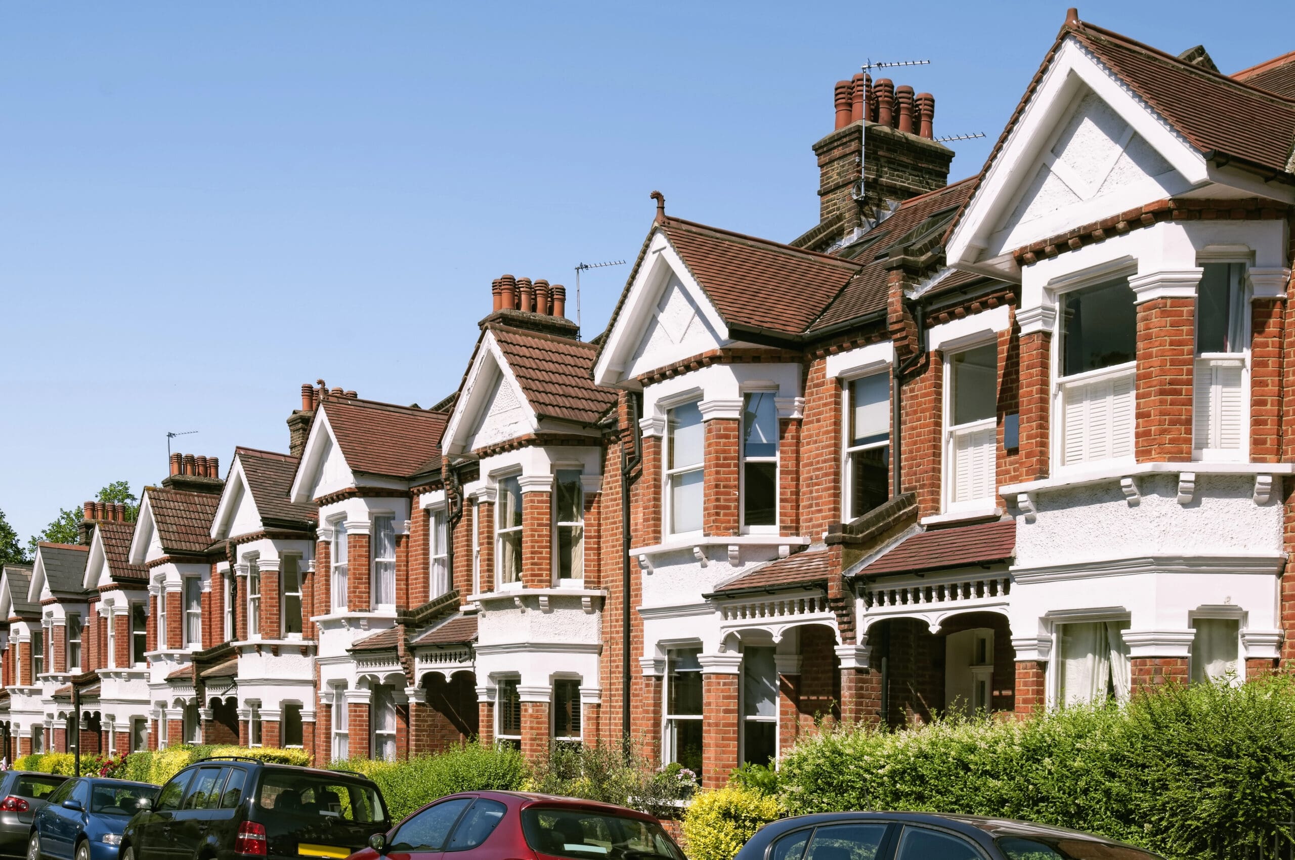 A row of traditional British terraced houses with red brick exteriors and white trims under a clear blue sky. Cars are parked along the street in front of the houses, and lush green hedges border the sidewalks.