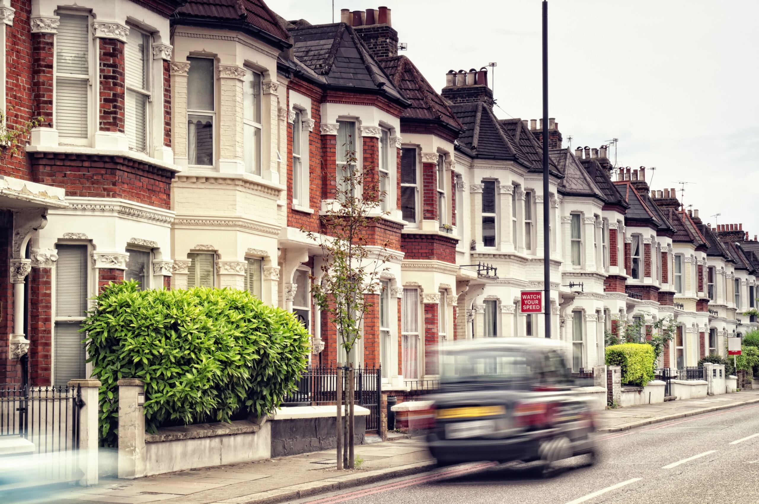 A row of Victorian-style terraced houses with red brick and white facades lining a street. A blurred black cab drives past, indicating motion. In front of the houses are small garden areas with neatly trimmed bushes and young trees.