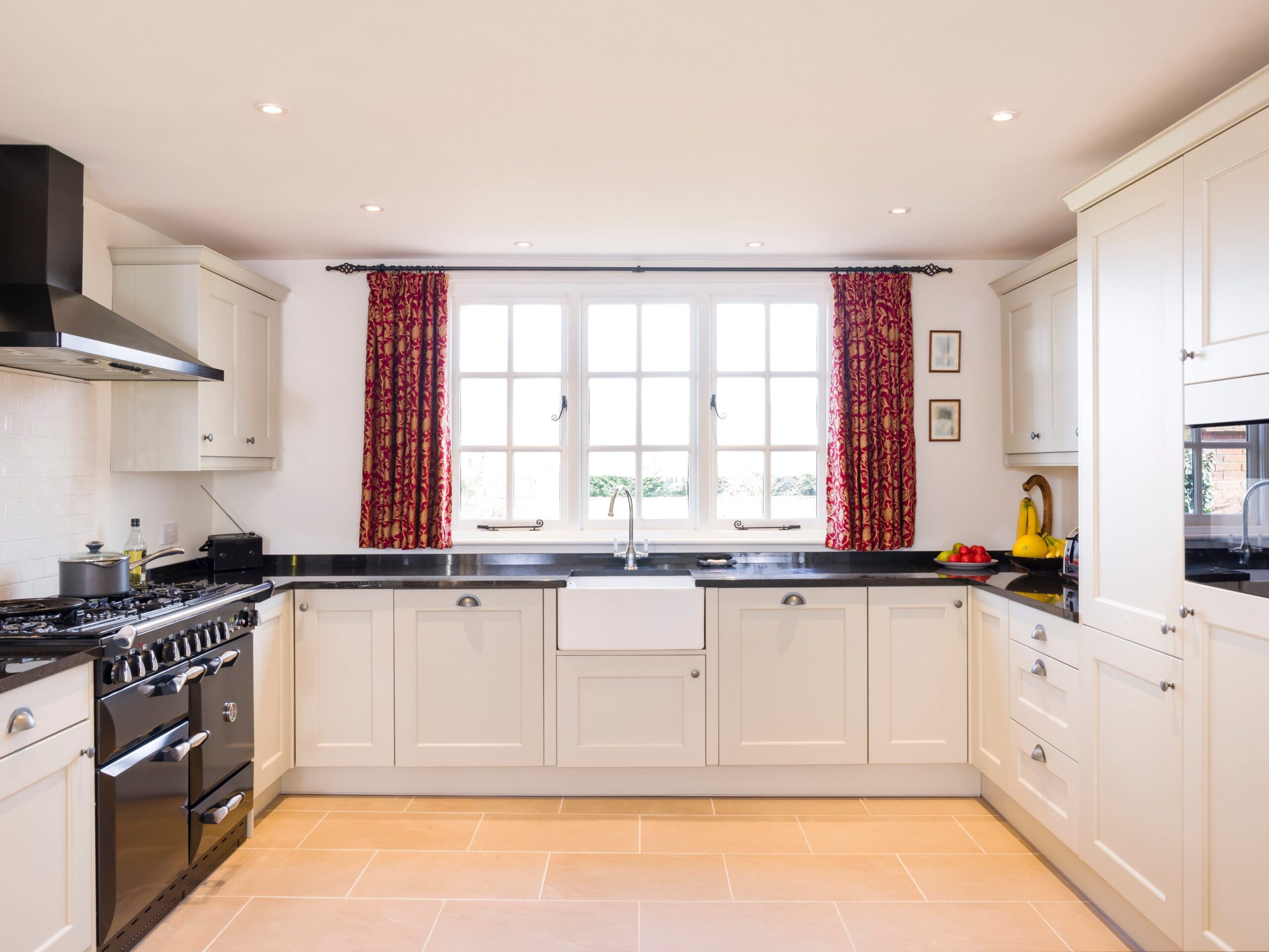 A bright, modern kitchen with white cabinetry, black countertops, and red patterned curtains on a large window. The room features a farmhouse sink, stainless steel appliances, and a tiled floor in a beige hue.