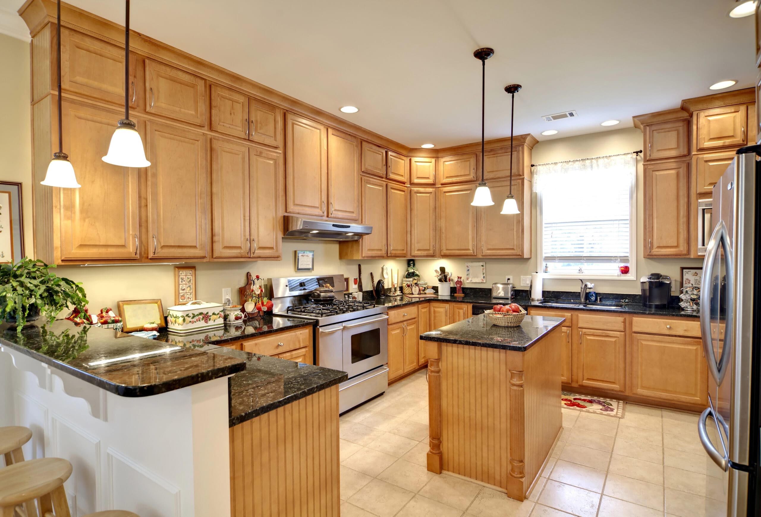 A spacious kitchen featuring wooden cabinets, stainless steel appliances, and granite countertops. An island with a granite surface stands in the center, surrounded by pendant lights. The room has tiled flooring and a large window with a white curtain above the sink.