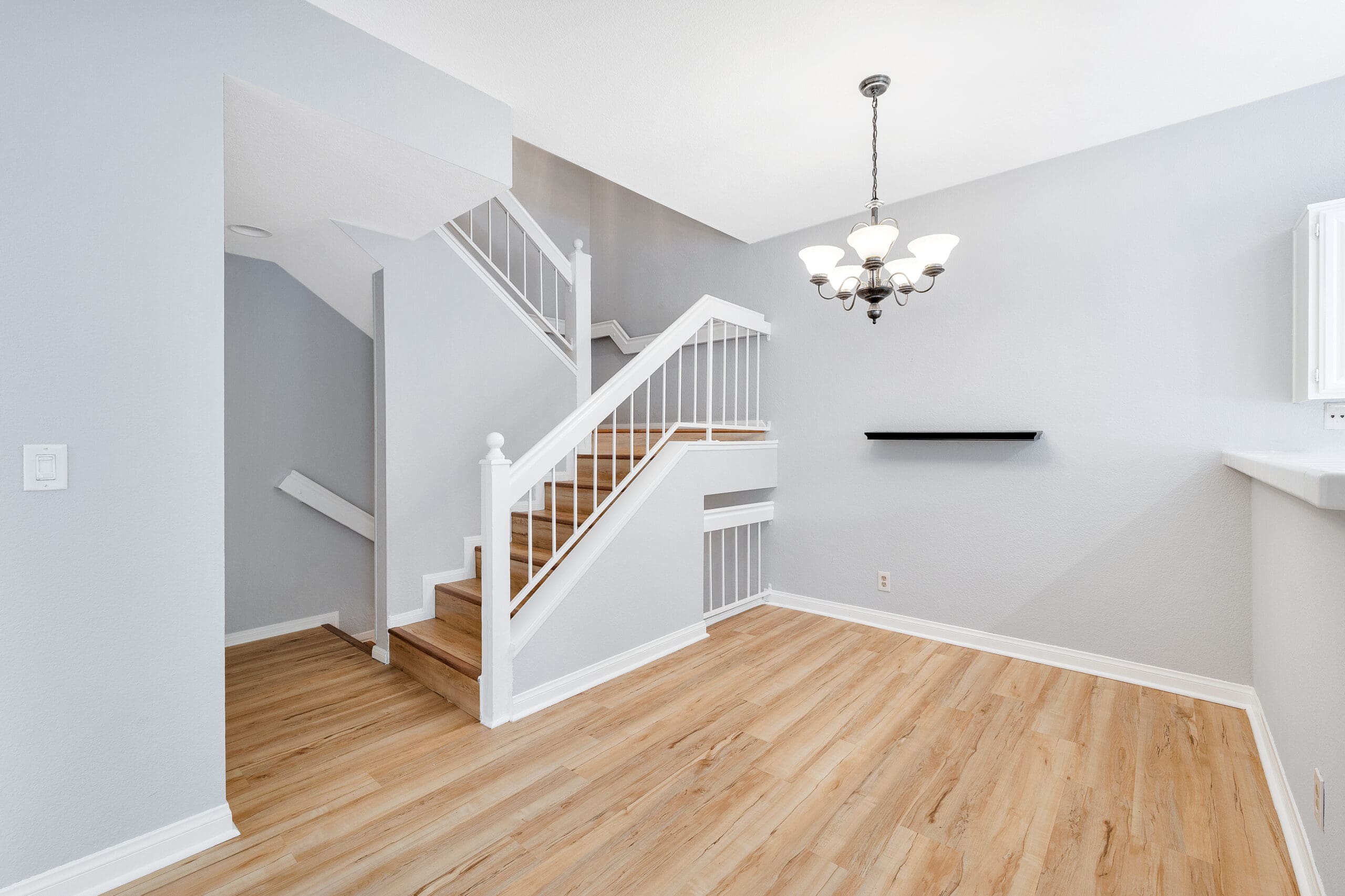 A modern, light-filled interior featuring a staircase with white railings and wooden steps. The room has light hardwood flooring, grey walls, and a contemporary chandelier hanging from the ceiling. A small black shelf is mounted on the wall under the chandelier.