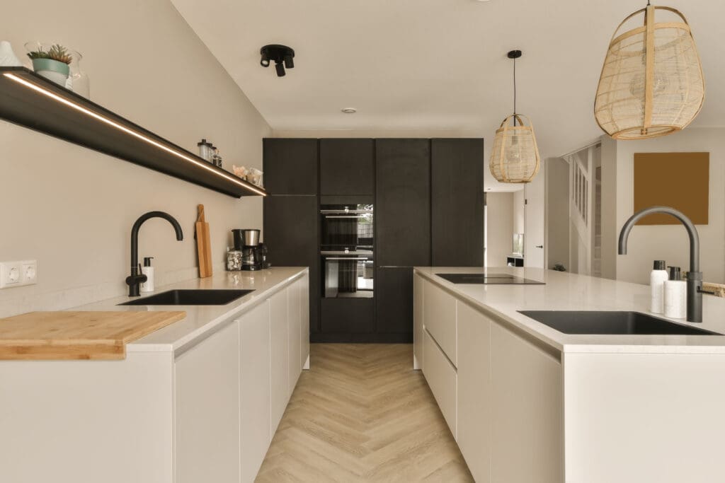 A modern, minimalist kitchen featuring sleek white cabinetry, a dual sink setup, black fixtures, and a built-in oven. Two pendant lights with wicker shades hang over the central island. A wooden cutting board and potted plant add a touch of warmth to the space.