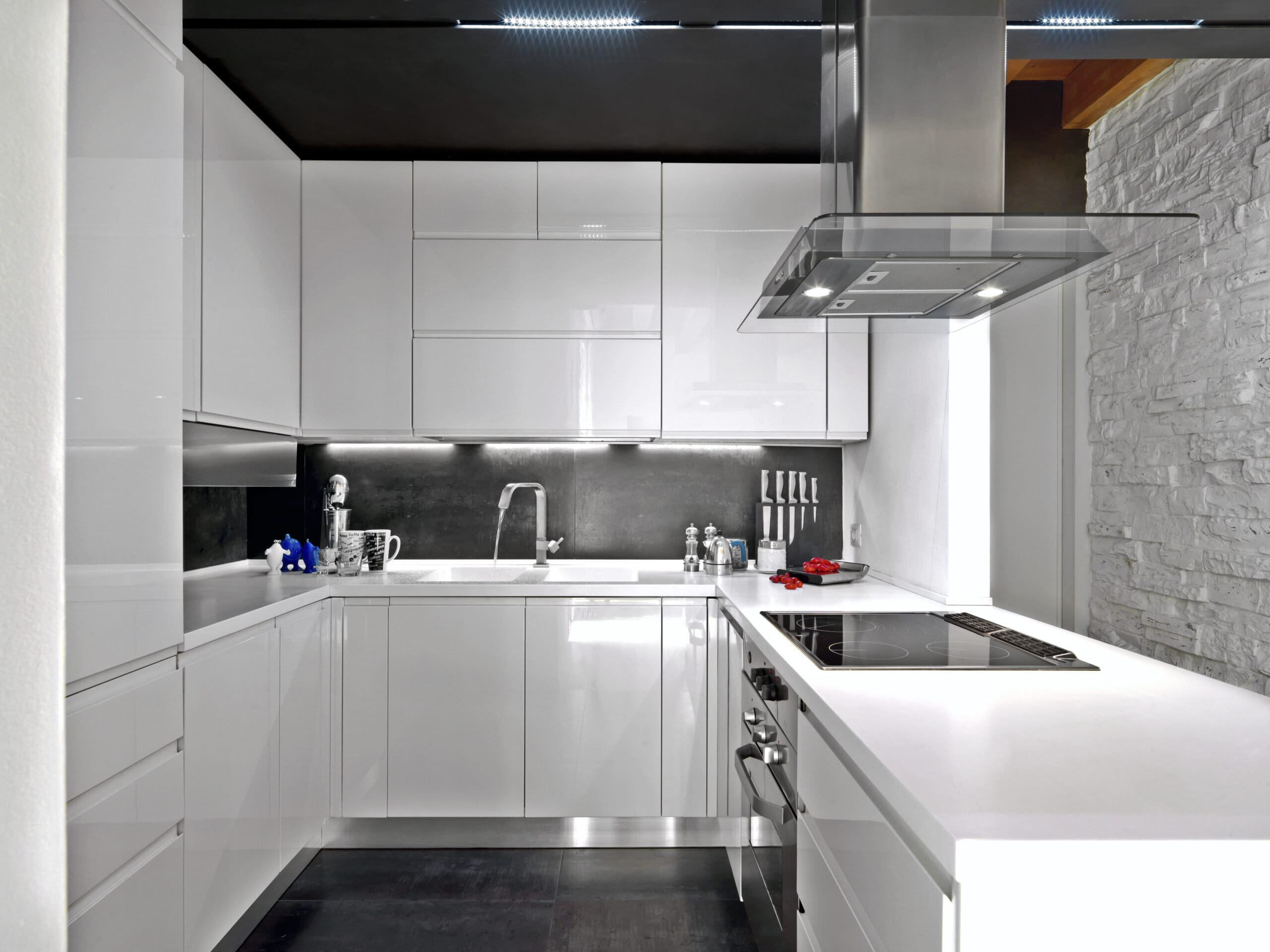 Modern kitchen with sleek white cabinetry and countertops. The kitchen features a built-in oven, electric cooktop, and stainless steel range hood. The black backsplash contrasts with the white surfaces. Kitchen utensils and appliances are neatly organized.