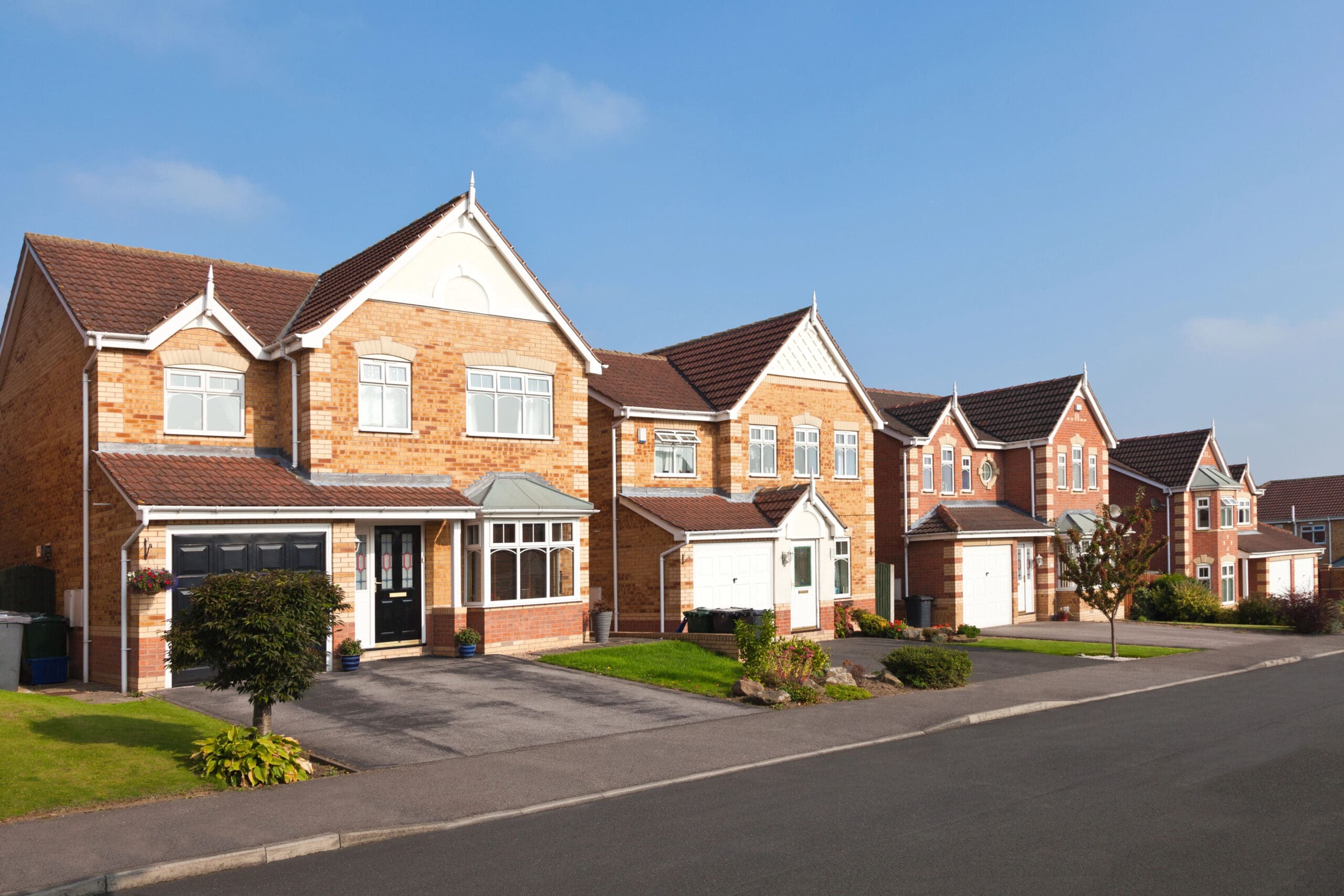 A row of modern suburban brick houses with gabled roofs, black and white front doors, and attached garages. The homes have well-kept lawns and are situated along a quiet street under a clear blue sky.