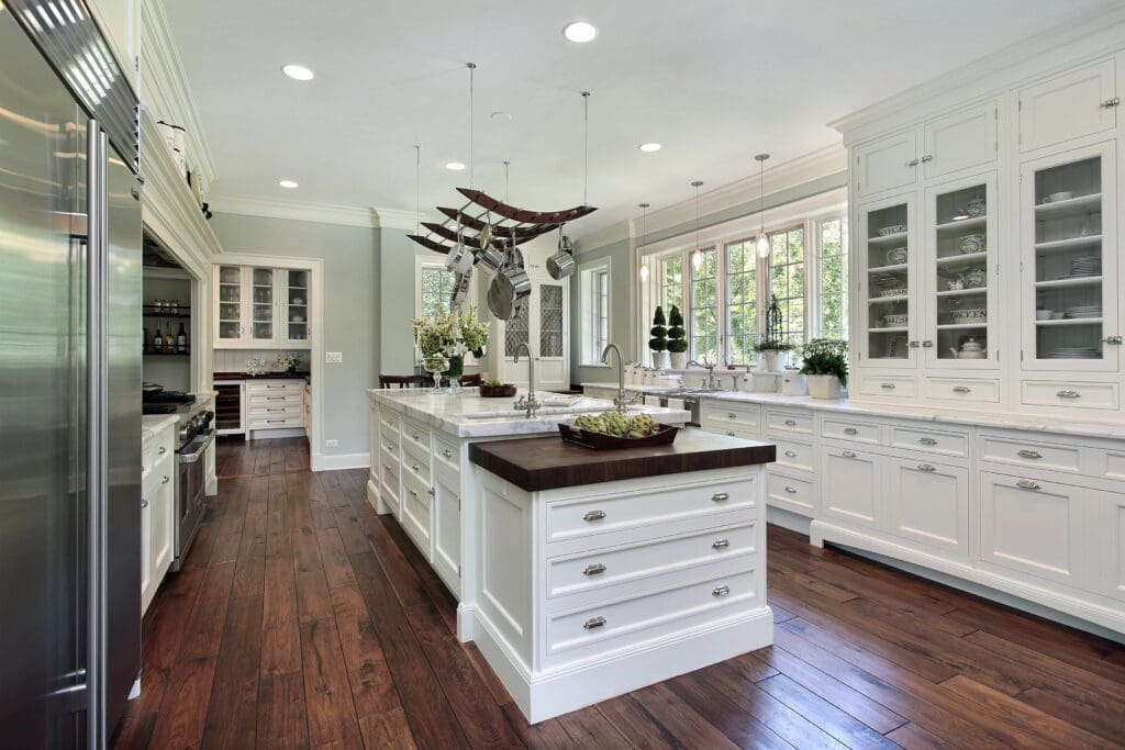 A modern kitchen featuring white cabinetry, a dark wood island with a hanging pot rack, and stainless steel appliances. The space is illuminated by recessed lighting and large windows that provide ample natural light. The wooden floors add warmth to the room.