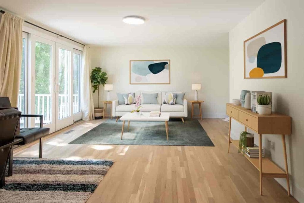 Bright living room with light wood floors, a white sofa with pastel cushions, a coffee table, large windows with curtains, modern artwork on the walls, houseplants, and a side console with decor items.