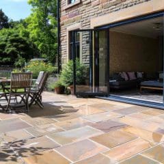 A sunny patio with stone tiles, a wooden table and chairs, and potted plants, next to a house with large glass doors open to a cozy living room. Lush green trees and plants surround the outdoor space.