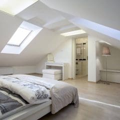 A modern attic bedroom with slanted ceilings, large skylights, a double bed with neutral bedding, a white vanity, a beige floor lamp, and light wood flooring. The room is bright and minimalist.