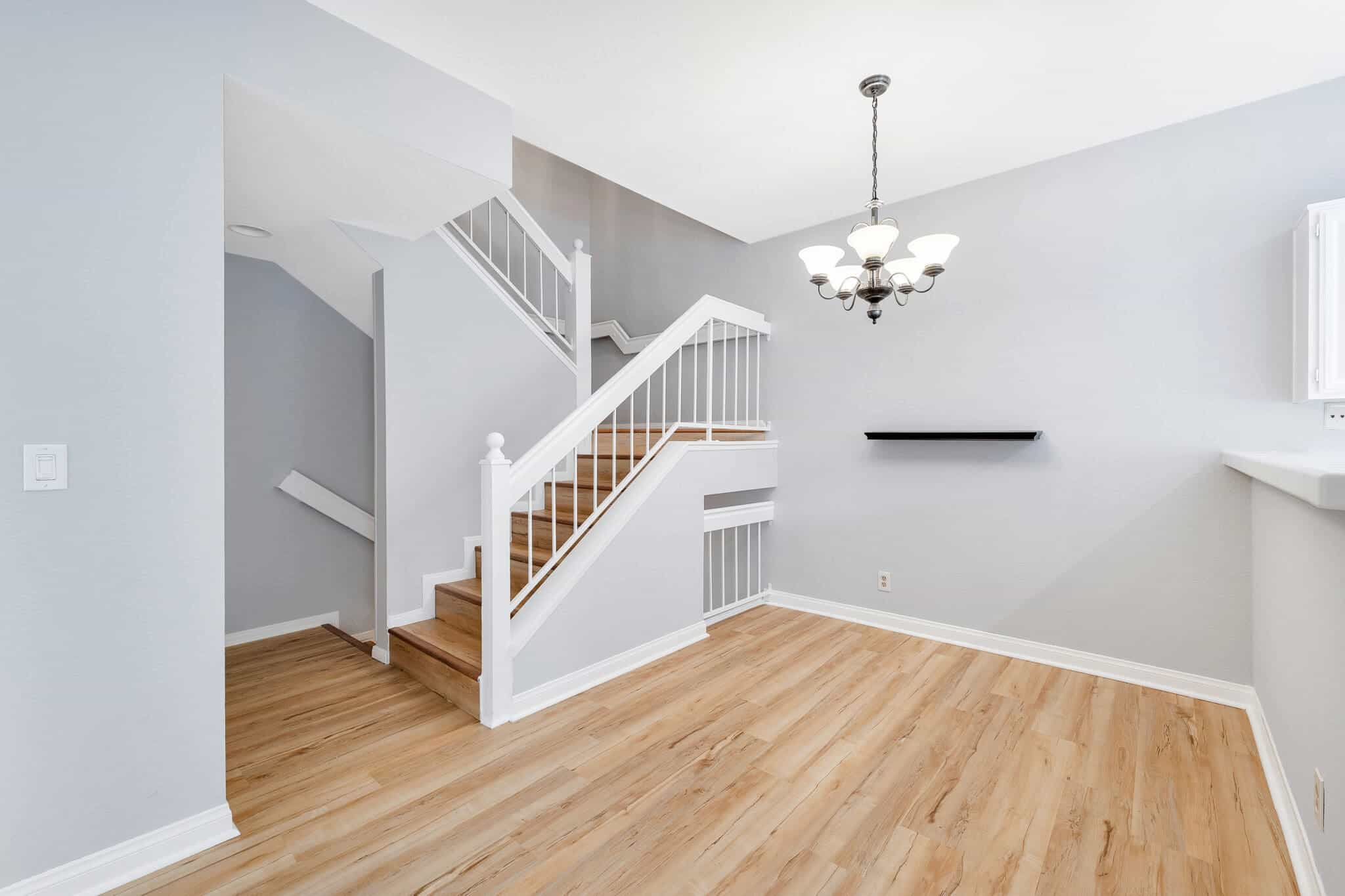 Modern, empty interior room with light wood flooring, a white staircase with wooden steps and white railing, light gray walls, and a chandelier hanging from the ceiling. A small black wall shelf is mounted on the wall.
